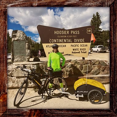 BB Day32 Photo Photo, Day 32 of the TransAmerica Bicycle Trail, Mandy with her bicycle in front of large sign that says: "Hoosier Pass, Elevation 11,539 feet, Continental Divide." There's also a smaller plaque that says: "Hoosier Pass across Continental Divide between South Park and the Blue River. Altitude 11,42 feet. Crossed by Fremont 1844. Named for Hoosier Gulch, discovered and worked as a placer camp by Indiana men 1860."