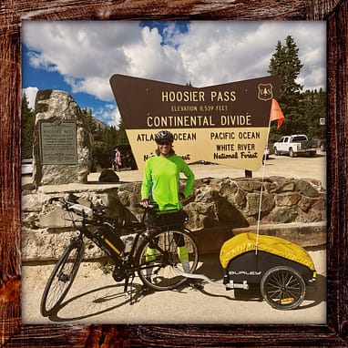 Photo, Day 32 of the TransAmerica Bicycle Trail, Mandy with her bicycle in front of large sign that says: "Hoosier Pass, Elevation 11,539 feet, Continental Divide." There's also a smaller plaque that says: "Hoosier Pass across Continental Divide between South Park and the Blue River. Altitude 11,42 feet. Crossed by Fremont 1844. Named for Hoosier Gulch, discovered and worked as a placer camp by Indiana men 1860."