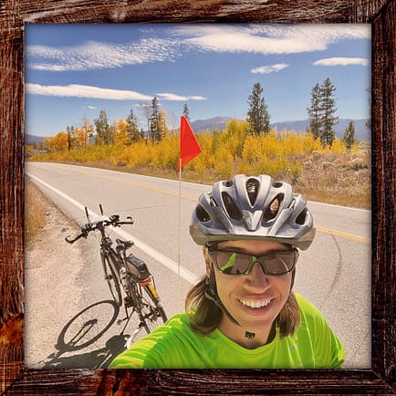 Photo, Day 29 of the TransAmerica Bicycle Trail, Mandy with her bicycle alongside a road lined with yellow aspen and mountains in the background
