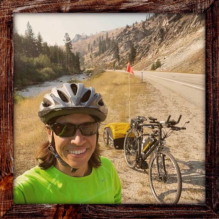 Photo, Day 16 of the TransAmerica Bicycle Trail, Mandy with her bicycle and trailer in front of mountains and a creek.