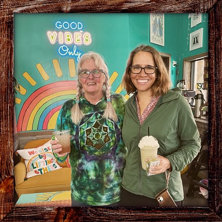 Photo, Day 26 of the TransAmerica Bicycle Trail, Mandy with Sally holding beverages at SunnyCup Ice Cream Shop in front of a sign with rainbow colors that says "Good Vibes Only"