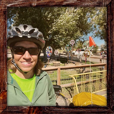 Photo, Day 27 of the TransAmerica Bicycle Trail, Mandy in front of park with windmills made of bicycle wheels and stained glass.