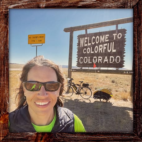 Photo, Day 28 of the TransAmerica Bicycle Trail, Mandy with her bicycle in front of a wooden sign "Welcome to Colorful Colorado"