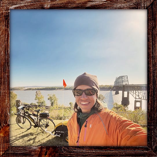 Photo, Day 46 of the TransAmerica Bicycle Trail, Mandy with her bicycle in front of the Mississippi River with a steel truss bridge in the background