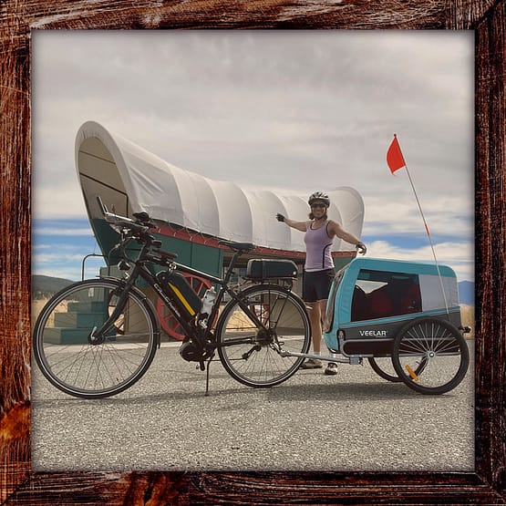 Photo, Day 08 of the TransAmerica Bicycle Trail, Mandy in front of covered wagon a pointing to her covered bicycle trailer, at a roadside historical marker for the Oregon Trail